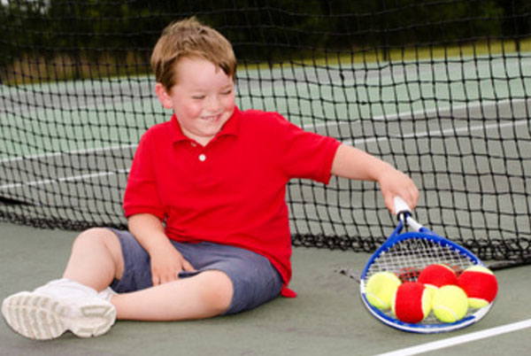 Мальчик сидит на земле возле сетки. Он держит теннисную ракетку, на которой пять мячиков